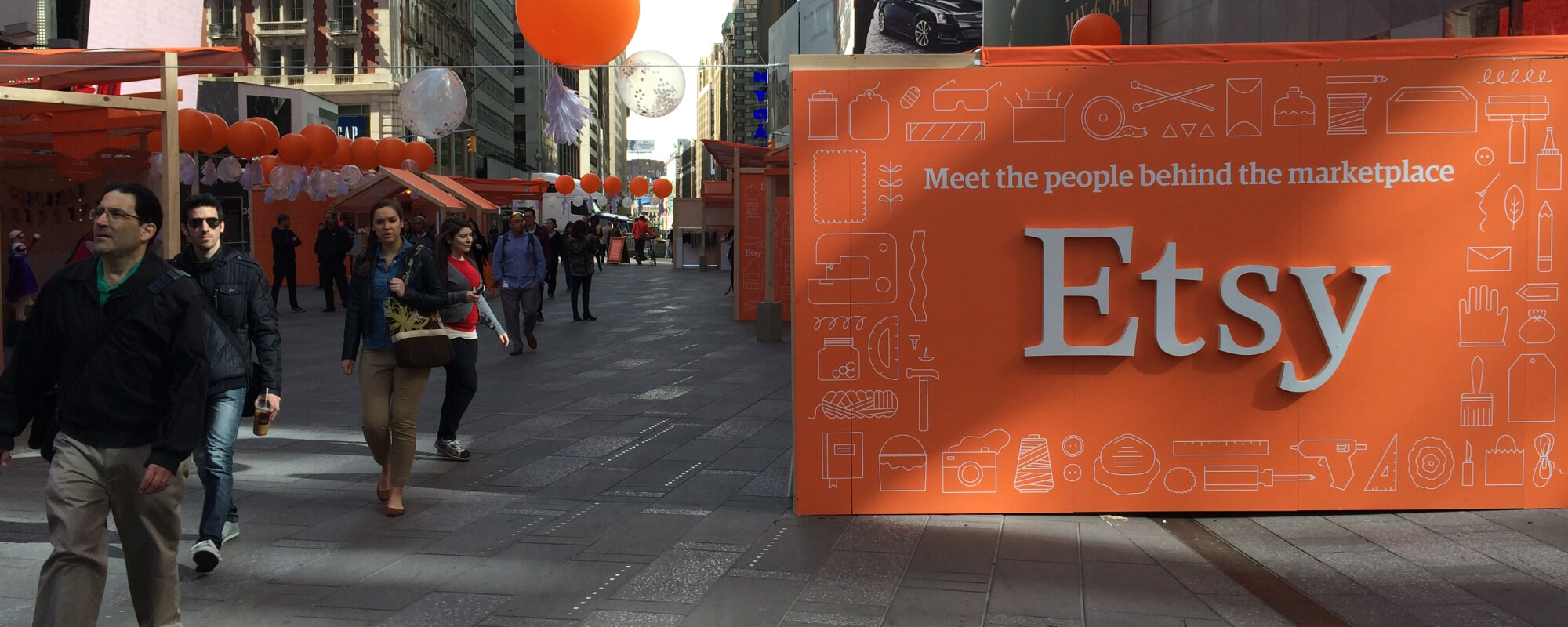 A group of people walking near a large orange sign that reads "Etsy"